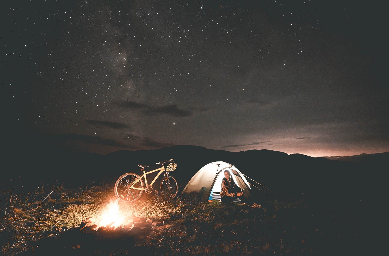 Young woman traveller having a rest at night camping near burning campfire, illuminated tourist tent, mountain bike under beautiful evening sky full of stars. Outdoor activity and tourism concept