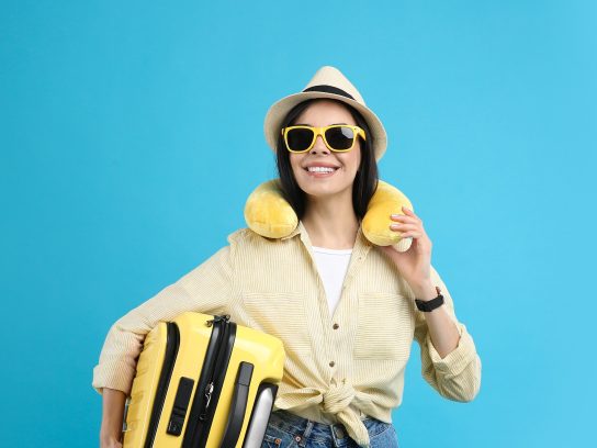 Happy female tourist with suitcase and travel pillow on light blue background-beste nekkussen vliegtuig