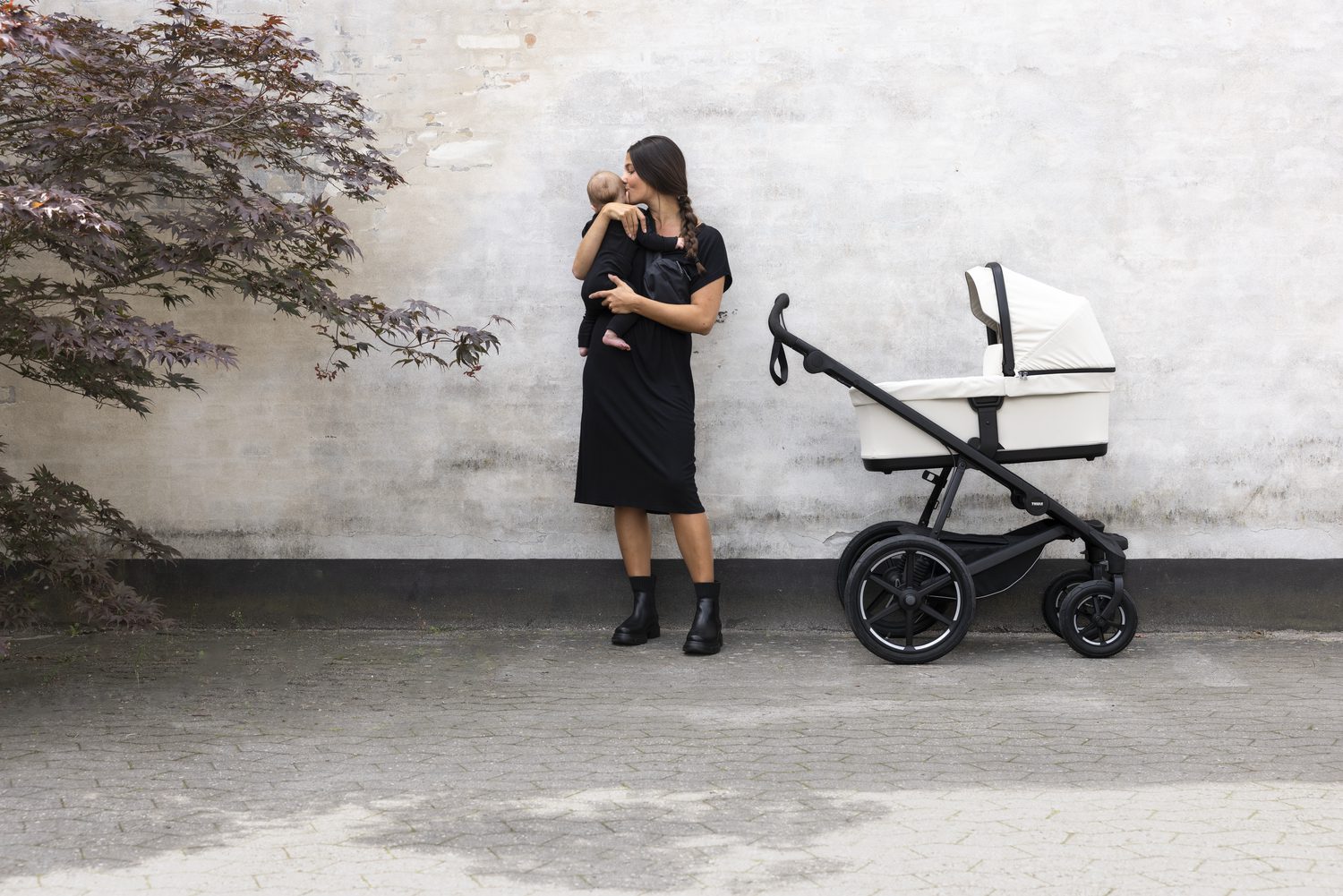 A woman holds and kisses a baby while standing beside a Thule Urban Glide 4-wheel stroller in color soft sand.