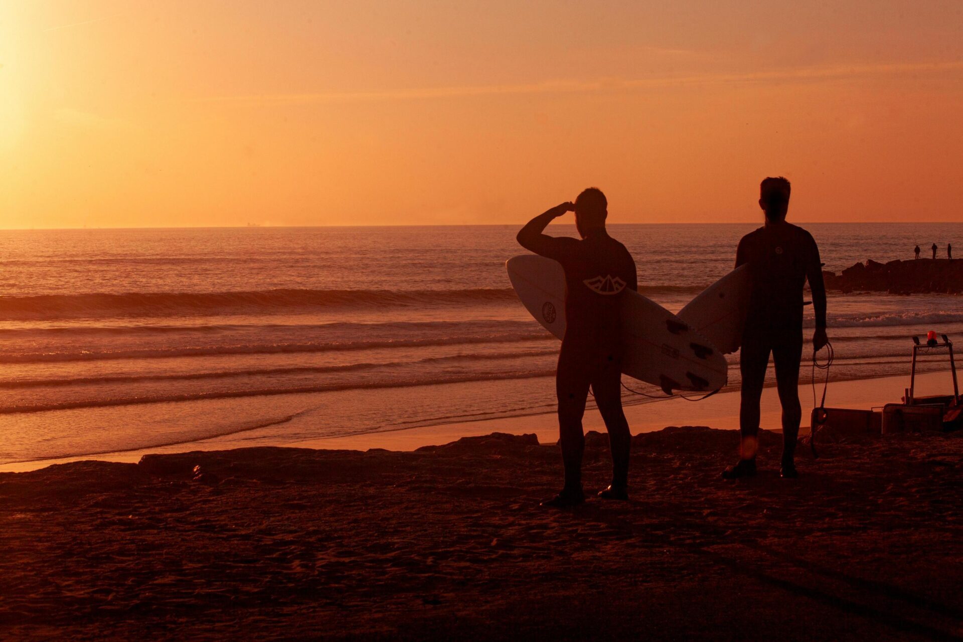 Twee personen met een surfboard aan de kust tijdens zonsondergang.