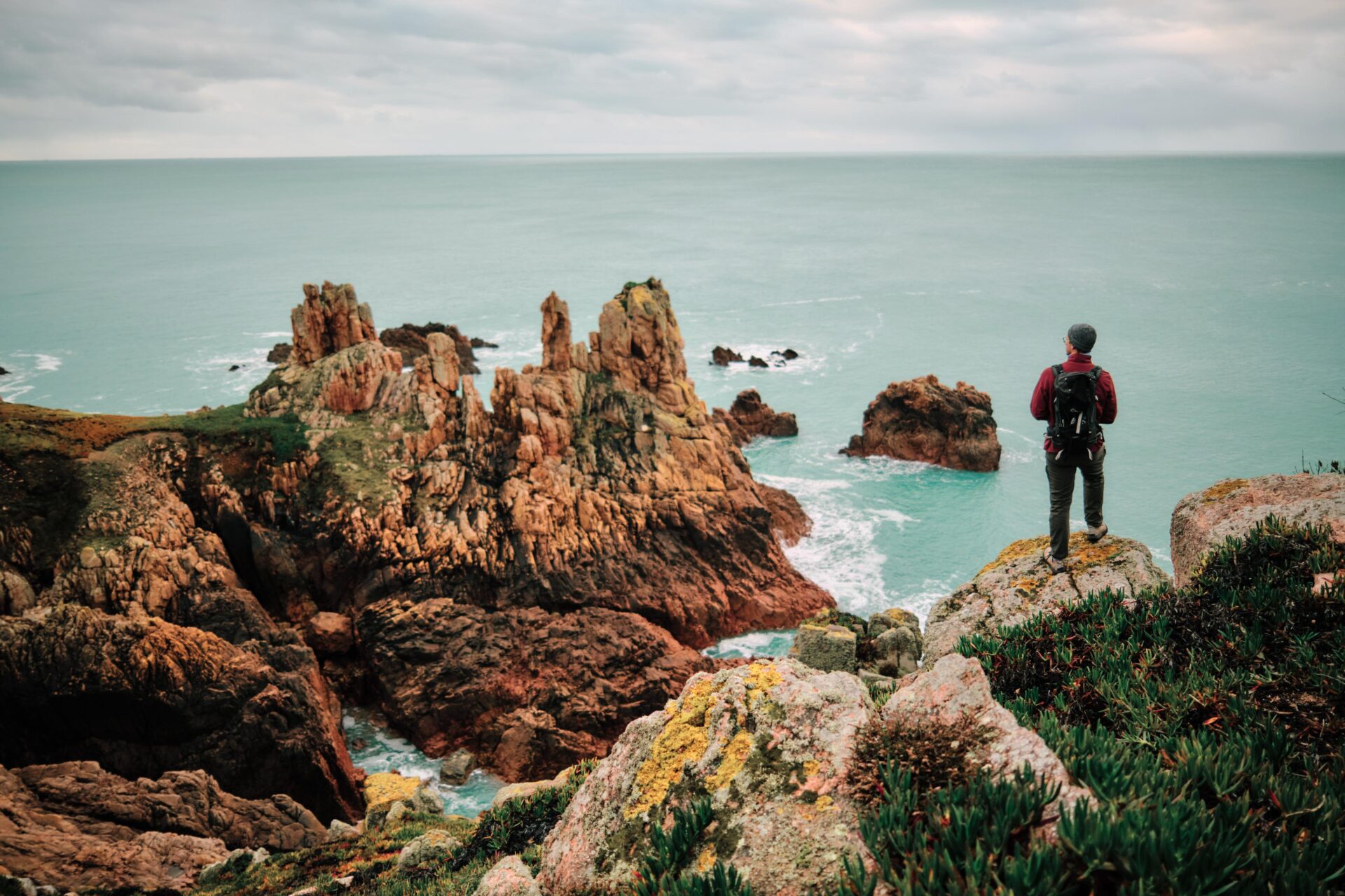 Een wandelaar staat aan de rand van een klif, waar rotsformaties uit de turquoise zee oprijzen langs de Jersey Tidal Trail.