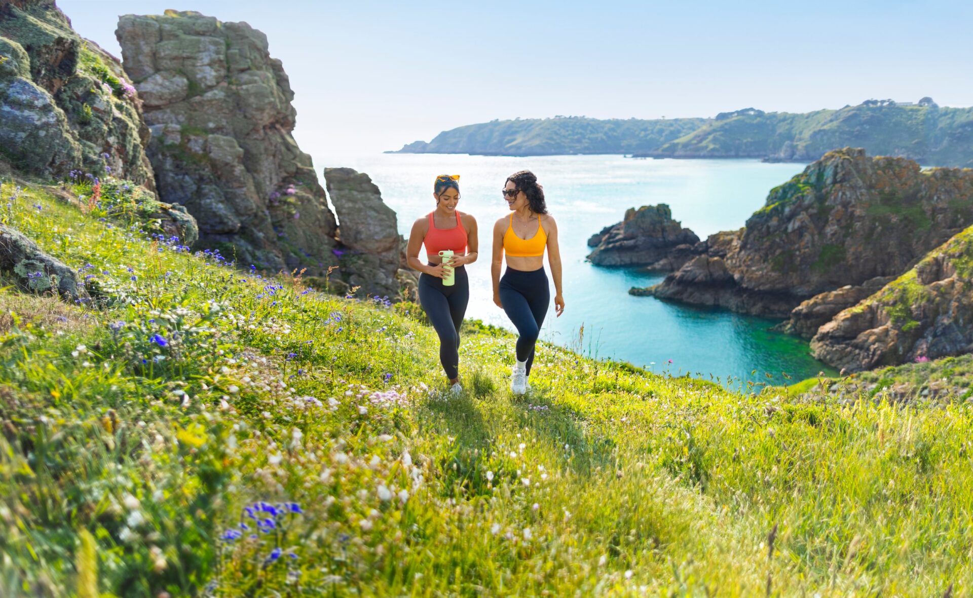 Twee mensen hiken door een grasveld met op de achtergrond het water en kliffen op Guernsey.